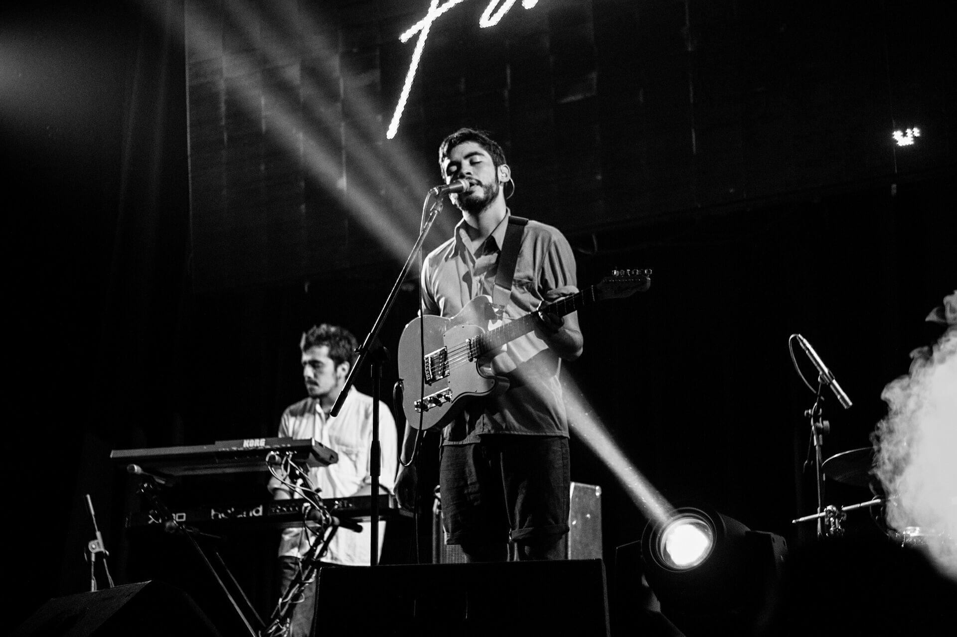 A black and white image of a band playing featuring a lead singer with a guitar and a drummer in the background.